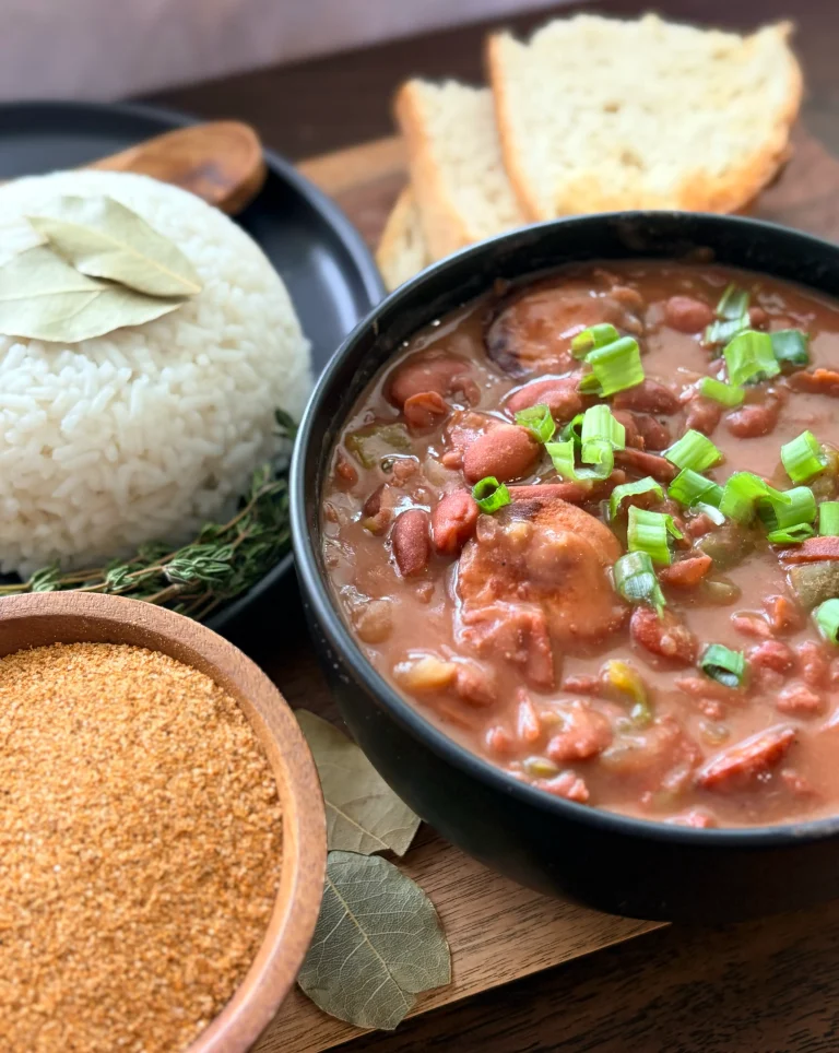 A bowl of red beans and rice with green onions sprinkled on top. There is a bowl of rice on the side with 3 pieces of fresh sourdough bread.