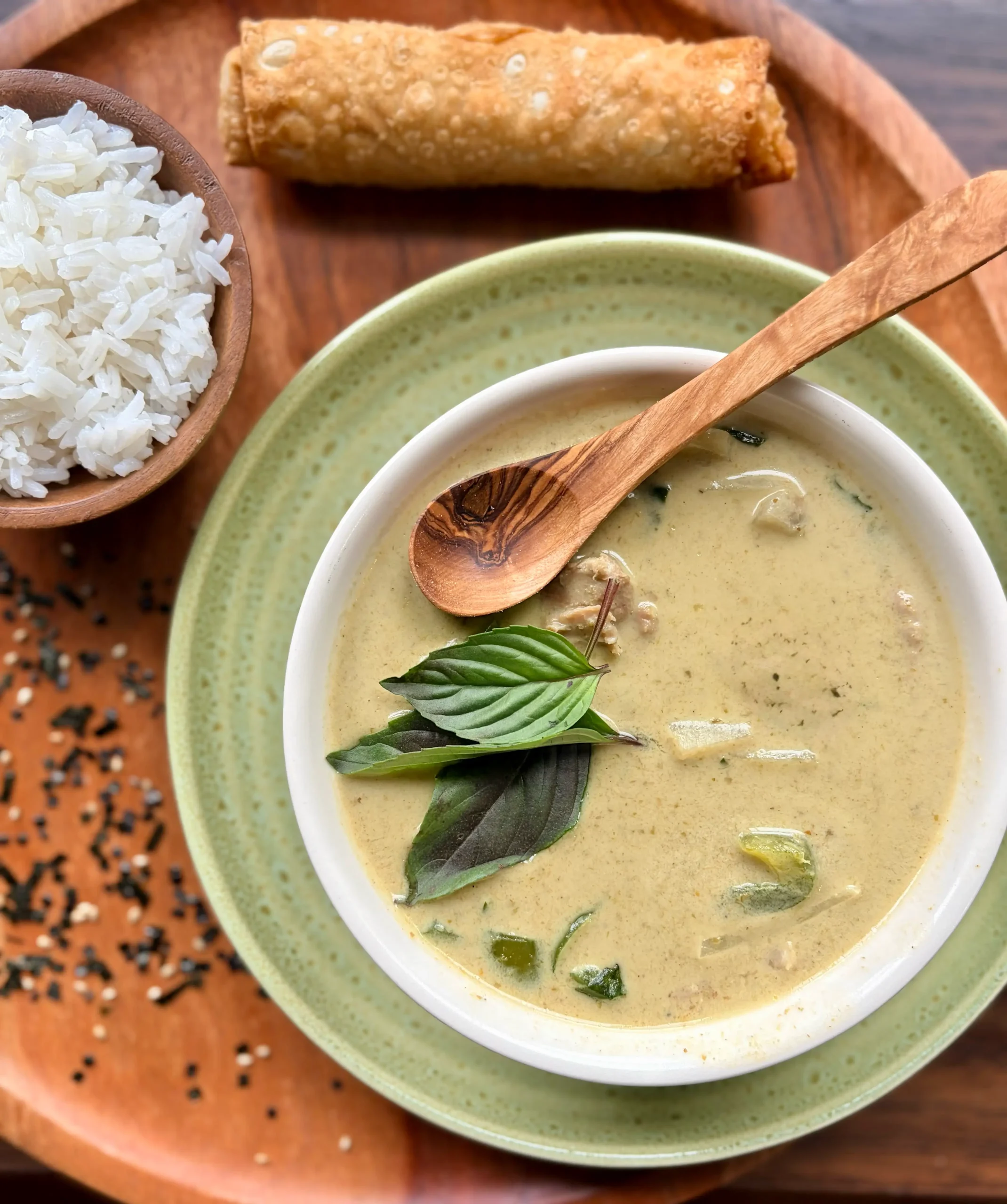 Bowl of thai green curry with a wooden spoon in the bowl. A side of white rice and an eggroll are next to the bowl.