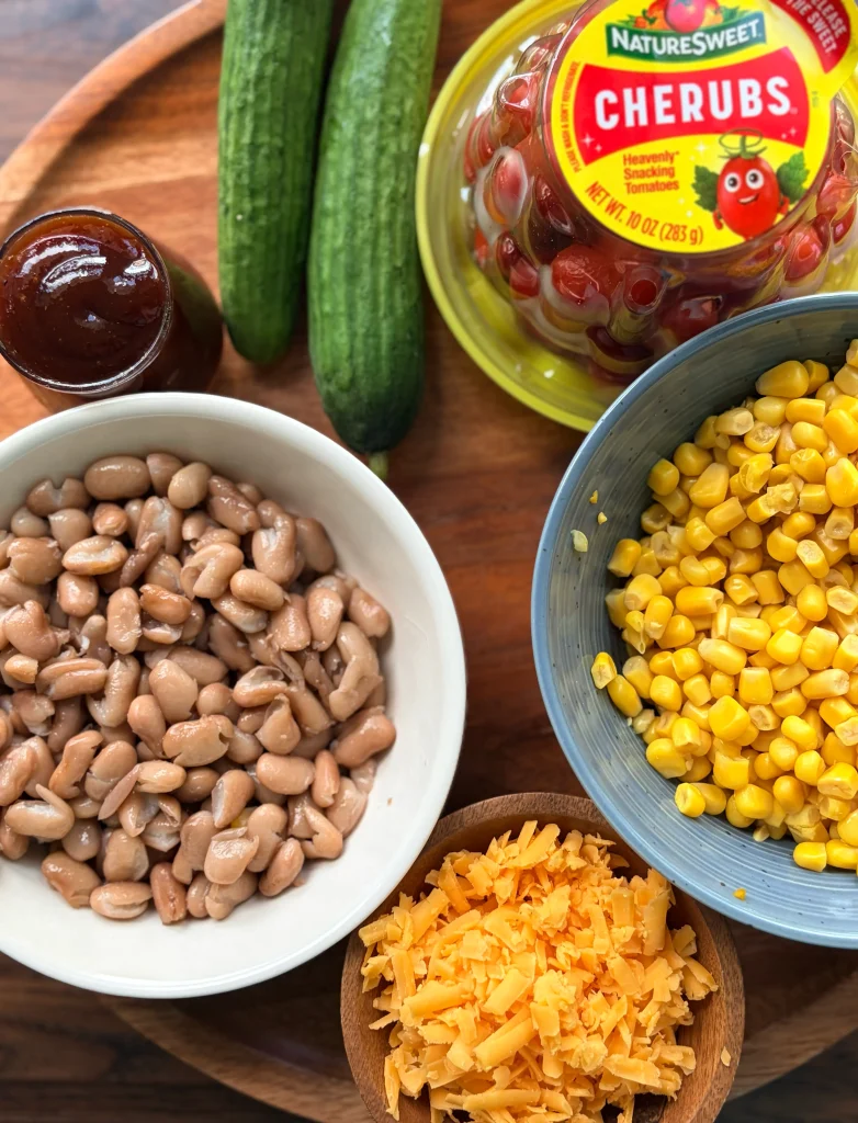 A bowl of pinto beans, 2 cucumbers, a bowl of corn, a package of cherry tomatoes and some shredded cheese.