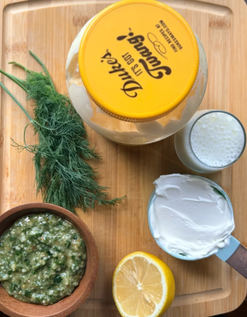 Ingredients for pesto ranch. Fresh pesto, mayo, sour cream, and buttermilk on a wooden cutting board.