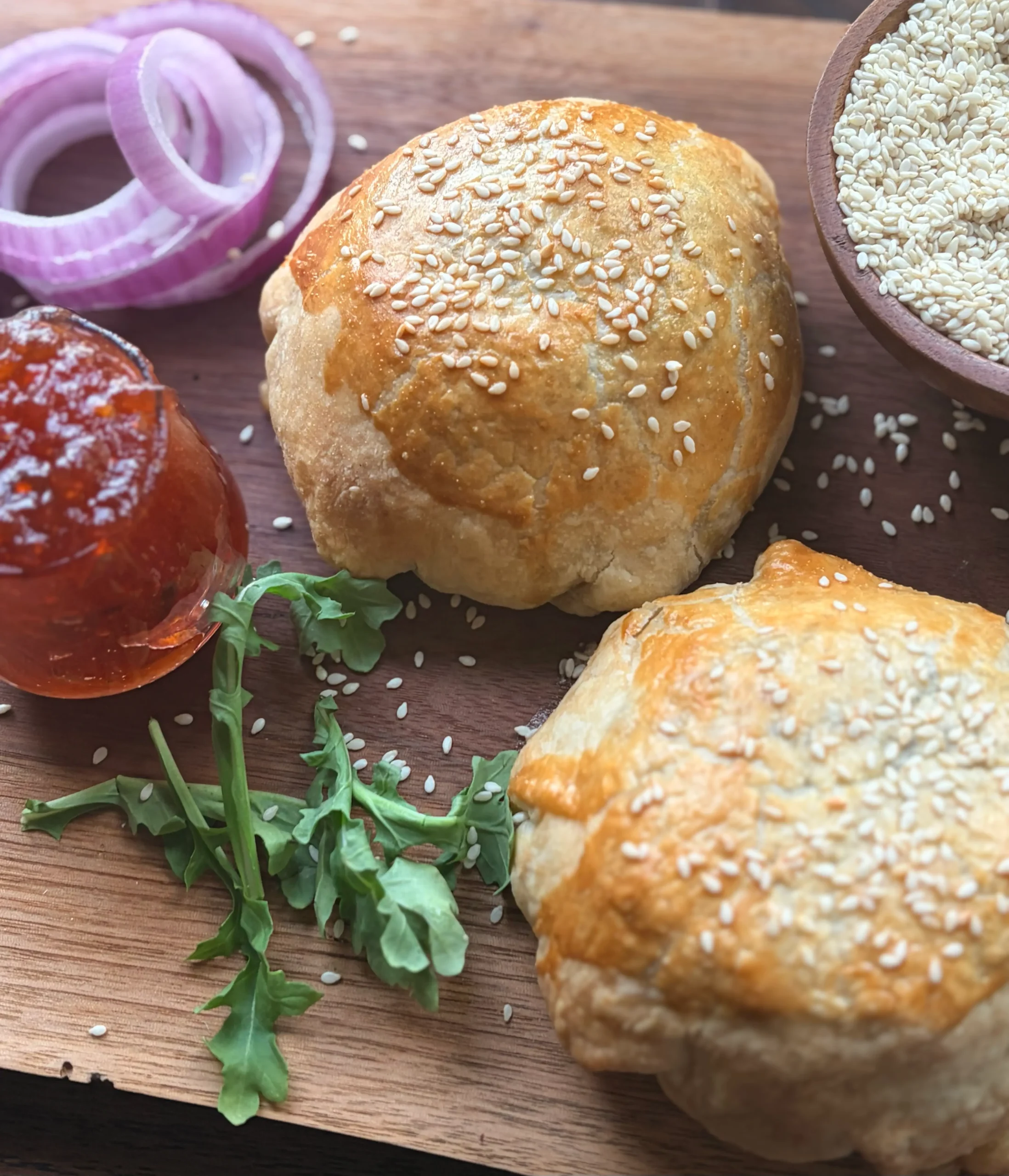 A jar of pepper jelly, red onion slices, and two burgers on a board.
