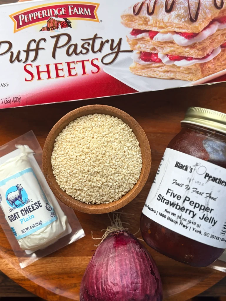 A package of puff pastry, a bowl of sesame seeds, a package of goat cheese, a jar of pepper jelly, and a red onion on a wooden board.