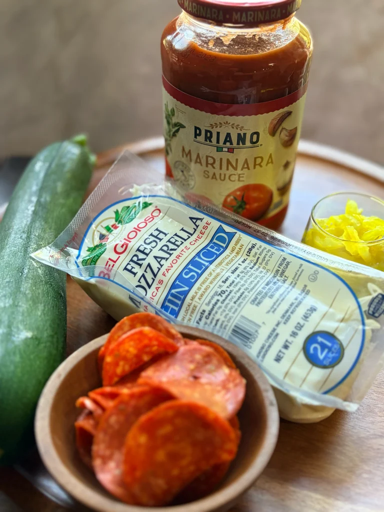 One zucchini with a bowl of pepperoni. A package of fresh mozzarella and marinara sauce on the side.