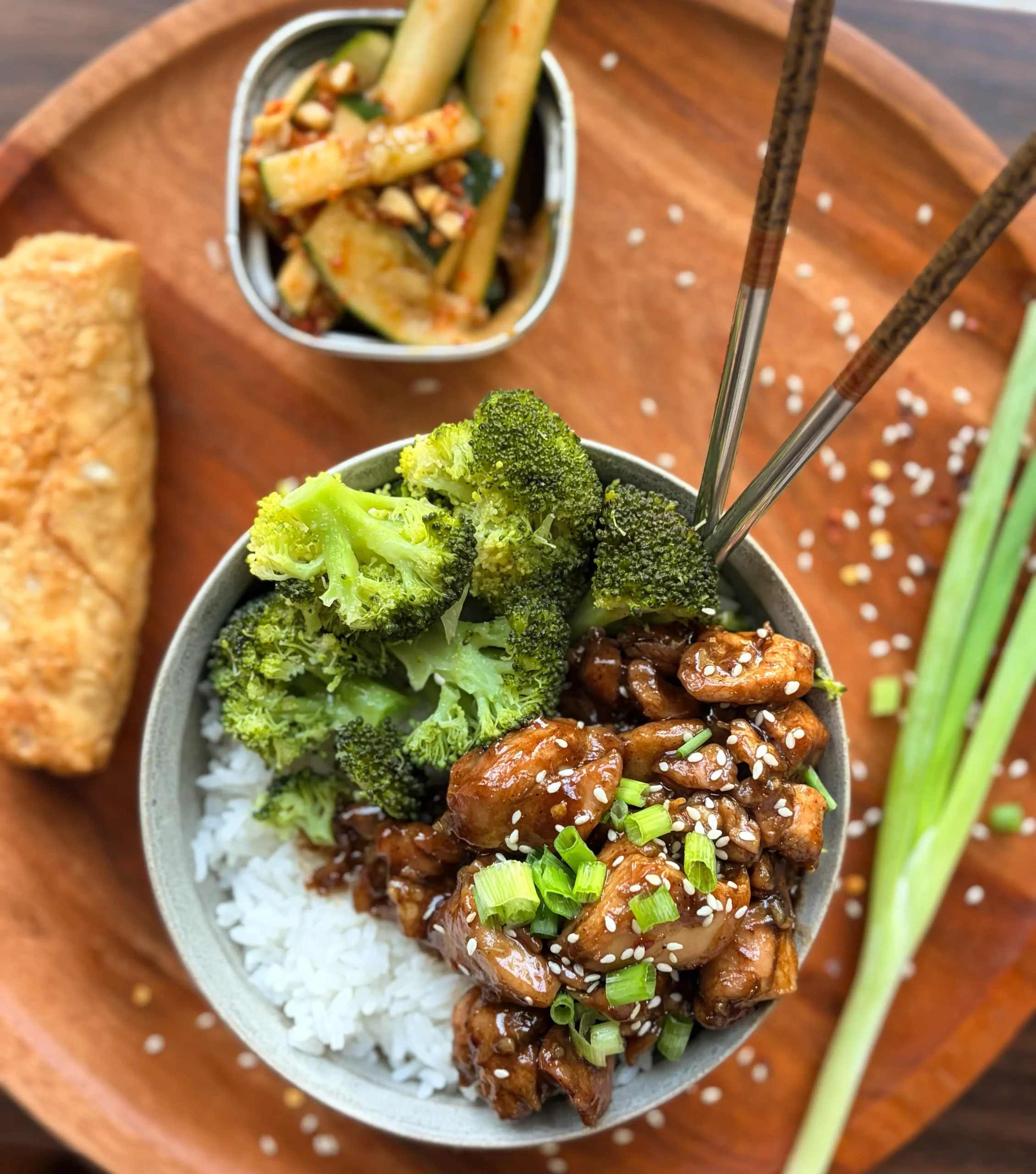 Honey-soy glazed chicken rice bowls with broccoli and an eggroll. Spicy cucumber salad on the side with a green onion sprig for garnish.